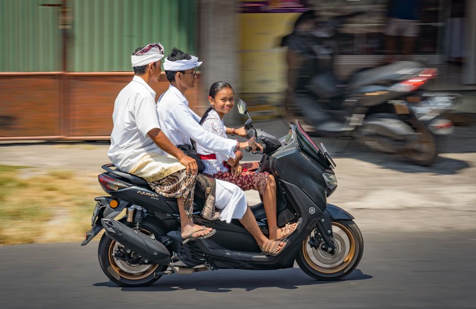 Balinese family in traditional attire riding a scooter on a sunny street.